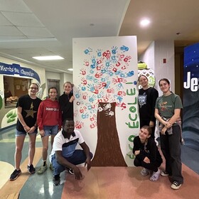 Un groupe de jeunes élèves et un adulte posent fièrement dans un corridor d'école autour d'une grande murale représentant un arbre fait d'empreintes de mains colorées, avec l'inscription « ÉCO-ÉCOLE ! ».