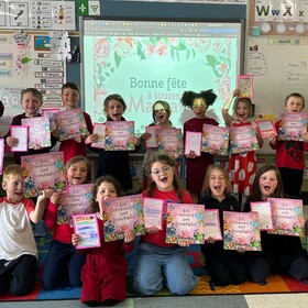 Une quinzaine de jeunes élèves souriants posent en classe avec leur enseignante, tenant fièrement des dessins et le livre « Les mamans sont formidables ! ».